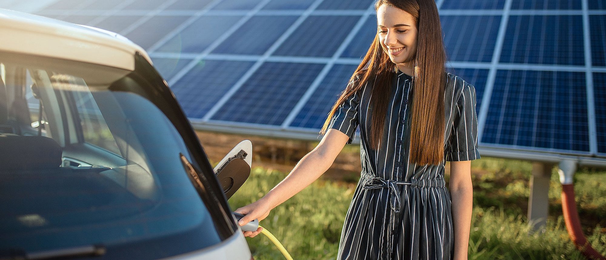 Lächelnde Frau lädt E-Auto und im Hintergrund ist eine große Photovoltaikanlage auf einer grünen Wiese